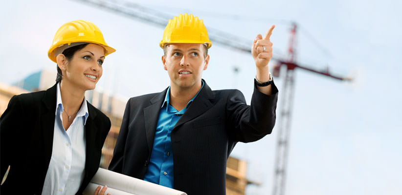 A man and a woman in business attire, holding blueprints, wearing hard hats, and drawing attention to something out of frame